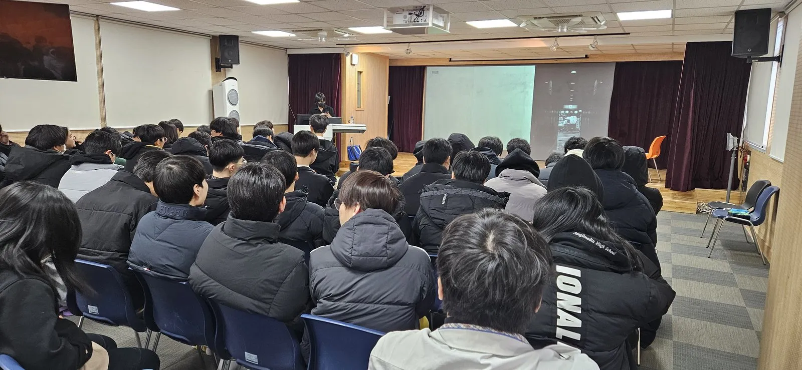 강당에 모인 학생들이 단상의 졸업생 강연을 듣는 장면