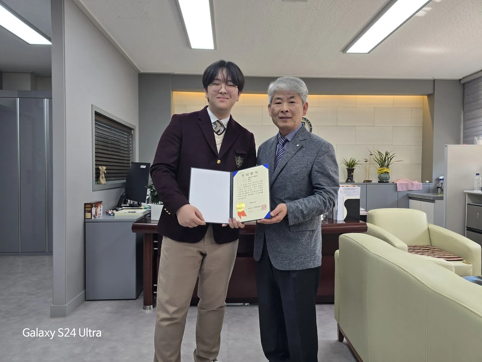 교장실에서 교장 선생님과 함께 장학증서를 펼쳐 든 학생